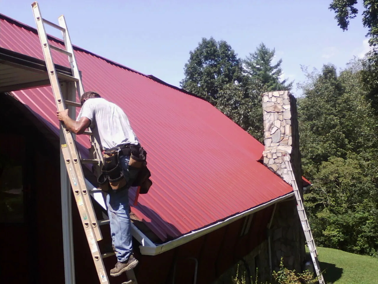 Roofer on a ladder beside a red metal roof and stone chimney — Soffit & Fascia Repair by Roofing Brothers in Wappingers Falls